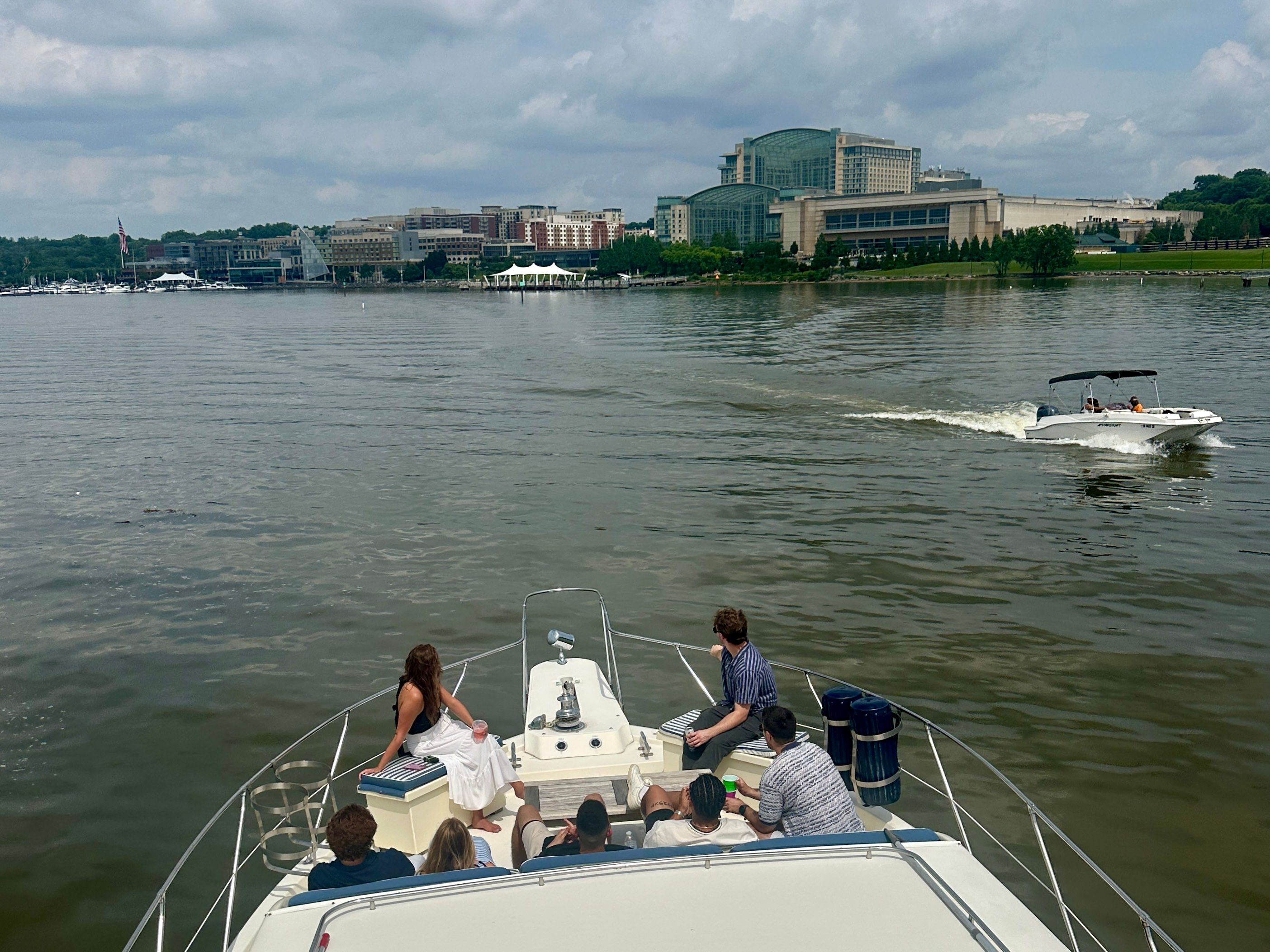 Group of people relaxing on a yacht bow cruising a calm river toward a city waterfront skyline, with a white speedboat passing nearby under a cloudy sky.