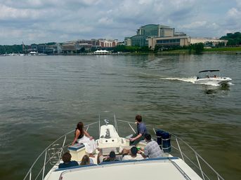 Group of people relaxing on a yacht bow cruising a calm river toward a city waterfront skyline, with a white speedboat passing nearby under a cloudy sky.
