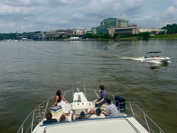 Group of people relaxing on a yacht bow cruising a calm river toward a city waterfront skyline, with a white speedboat passing nearby under a cloudy sky.