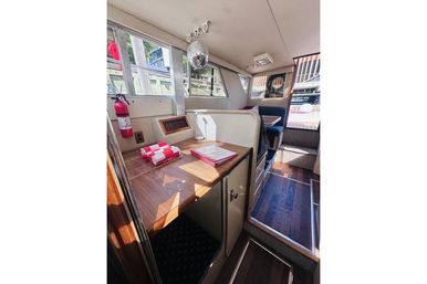 Sunlit boat cabin interior with wood table and cabinetry, navy dinette seating, folded red-and-white towels, quirky hanging disco ball, fire extinguisher, and windows overlooking a marina dock