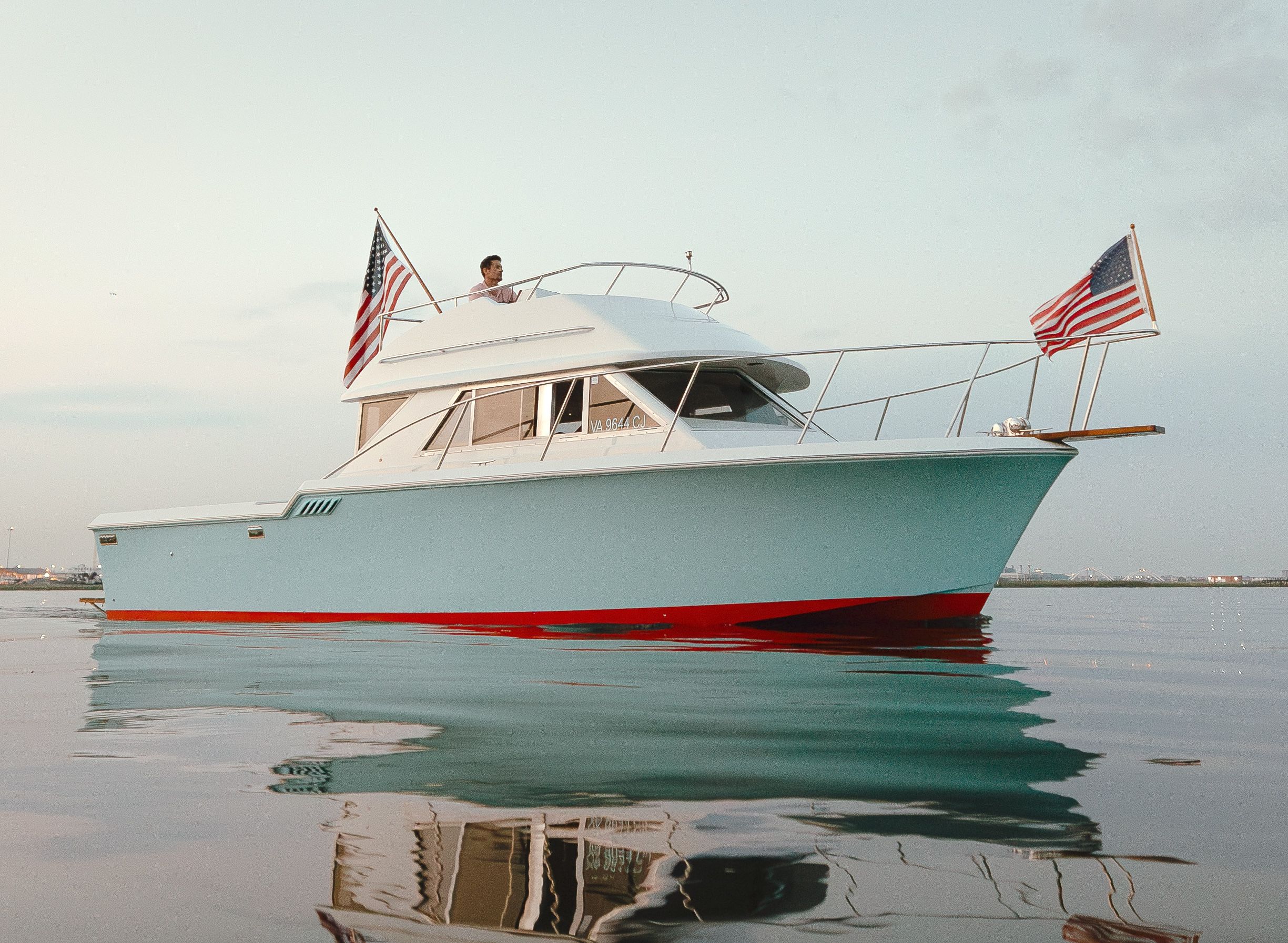 Light-blue motor yacht with a red waterline and American flags on bow and stern floating in a calm coastal harbor at dusk, crisp water reflection