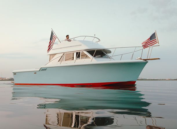 Light-blue motor yacht with a red waterline and American flags on bow and stern floating in a calm coastal harbor at dusk, crisp water reflection
