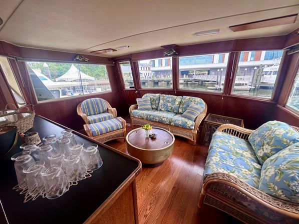 Sunlit yacht salon with wicker sofas and armchairs in tropical floral and striped cushions, round wooden coffee table, stacked glasses on a counter, hardwood floor and large windows overlooking a marina and waterfront buildings.