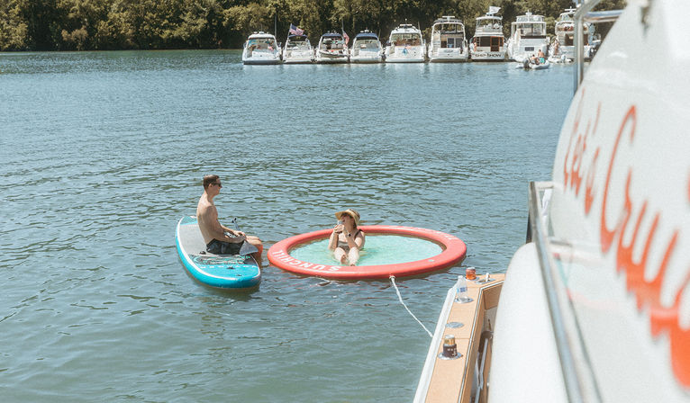 Sunny lake scene: man on a paddleboard chats with a woman lounging in a circular inflatable floating pool tethered to a nearby boat, with yachts docked along a tree-lined shore.