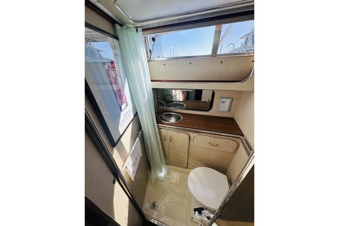 Cozy boat bathroom (marine head) with round stainless sink set in wood countertop, mirrored cabinet, shower curtain, compact toilet and porthole window showing sailboat masts
