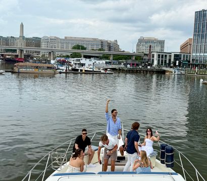 Group of friends enjoying the bow of a yacht at the Washington, D.C. waterfront marina, with waterfront buildings and the Washington Monument visible under a cloudy sky.