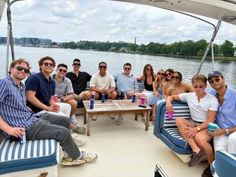 A dozen friends lounging on a striped-seated party boat, sipping drinks on a calm river with a tree-lined shore and distant city skyline under a cloudy summer sky.