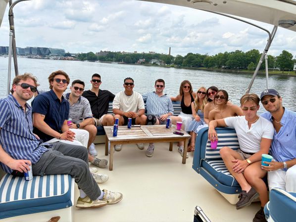 A dozen friends lounging on a striped-seated party boat, sipping drinks on a calm river with a tree-lined shore and distant city skyline under a cloudy summer sky.