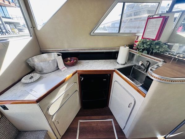 Cozy yacht galley with marble countertop, stainless sink, mini fridge, ice bucket and fruit bowl under porthole windows with marina view