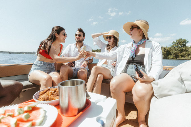 Four friends toasting drinks on a sunny lake boat trip, wearing summer hats and sunglasses with watermelon and snacks on a tray.
