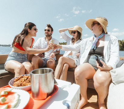 Four friends toasting drinks on a sunny lake boat trip, wearing summer hats and sunglasses with watermelon and snacks on a tray.