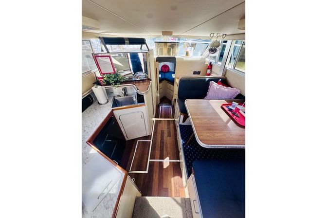 Cozy motorboat cabin interior with compact galley and sink, marble-look counters, warm wood plank flooring, navy dinette seating and wooden table, striped throw pillow and windows with waterfront views.