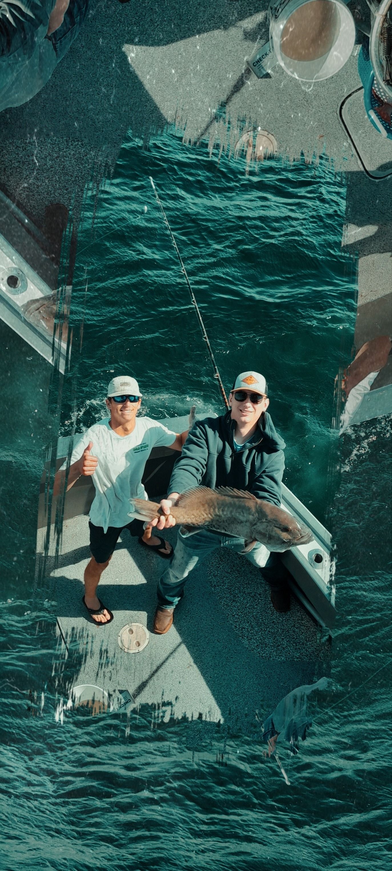 Two anglers on a sport-fishing boat proudly holding a large catch and giving a thumbs-up over teal ocean waves.