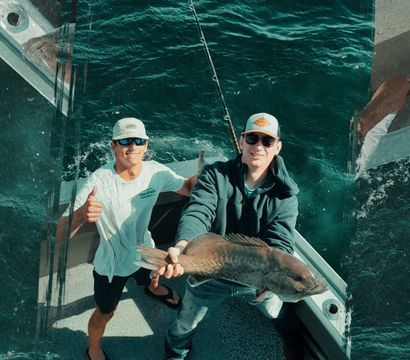 Two anglers on a sport-fishing boat proudly holding a large catch and giving a thumbs-up over teal ocean waves.