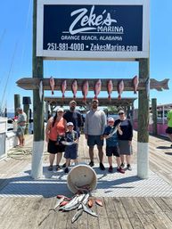 Smiling group on a sunny Orange Beach, Alabama marina dock with red snapper and other fish hung on a wooden rack and spilling from a bucket, boats and water in the background.