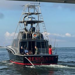 Rear view of a sportfishing boat with a tall observation tower, anglers and passengers on the stern holding multiple fishing rods, cruising in calm coastal waters under a clear blue sky.