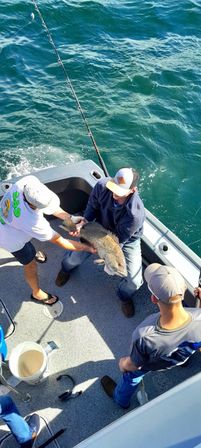 Three anglers on a small fishing boat holding a large catch over turquoise offshore waters, fishing rod, bucket, and boat deck visible