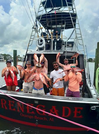Seven anglers on a charter fishing boat at an Alabama Gulf Coast marina celebrating while holding multiple red snapper under a cloudy sky.