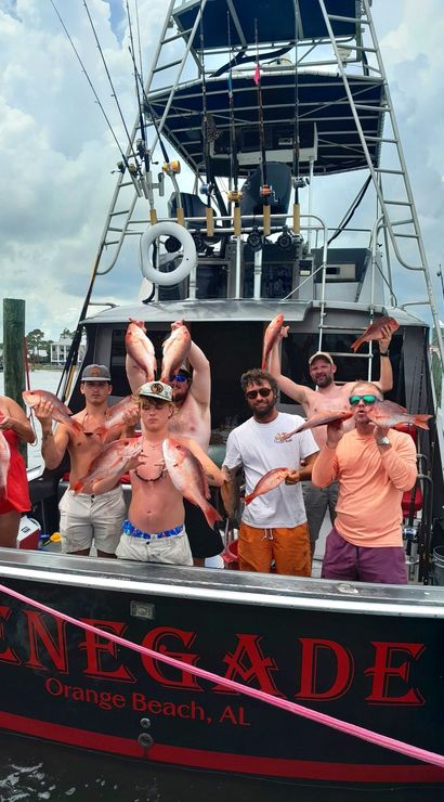 Seven anglers on a charter fishing boat at an Alabama Gulf Coast marina celebrating while holding multiple red snapper under a cloudy sky.