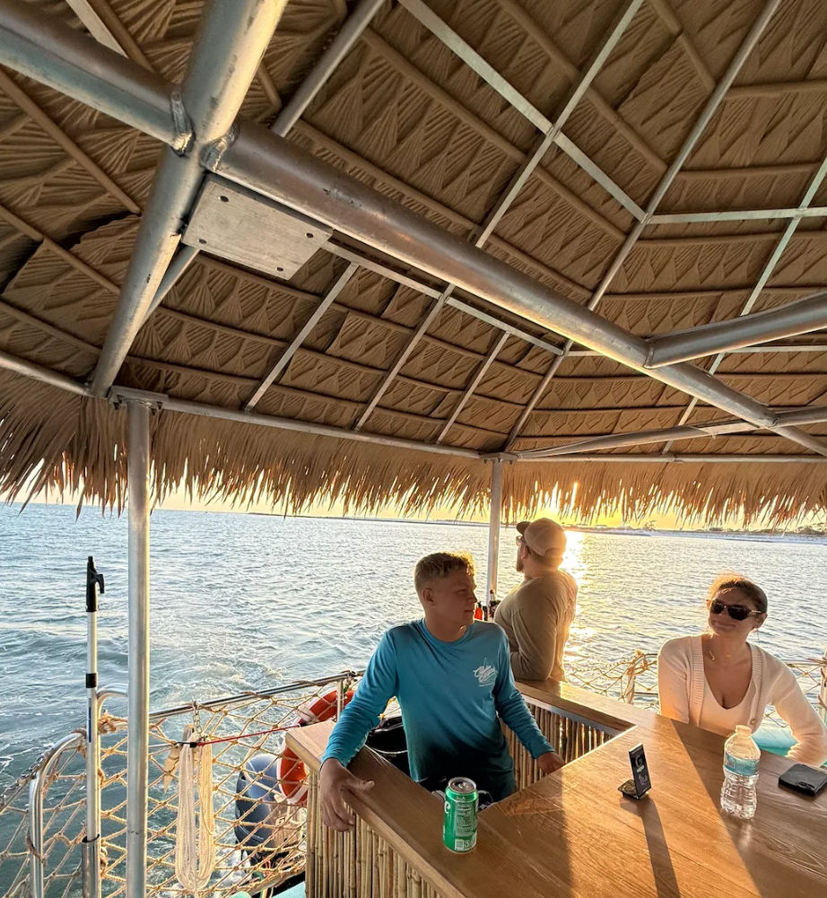Three people relaxing under a thatched tiki-roof on a boat at sunset, ocean view with warm golden glow and tropical cruise vibe