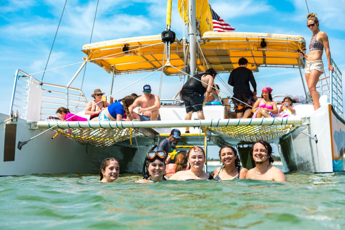 Smiling group of swimmers in green coastal water beside a yellow-canopied catamaran with people relaxing on deck and snorkeling gear, sunny day boat outing