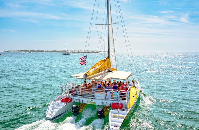 Bright yellow catamaran cruise with passengers and an American flag sailing on sparkling blue coastal waters under a sunny sky.