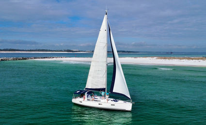 White-sailed yacht gliding on turquoise coastal waters near a white-sand beach and low shoreline under a partly cloudy blue sky, with a few people on deck.