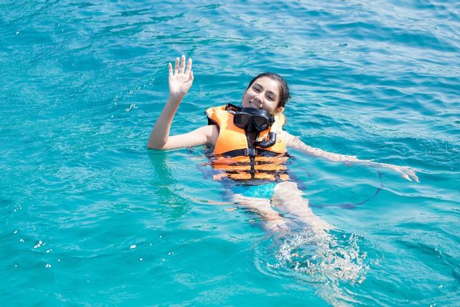 Smiling person in an orange life jacket and snorkel mask floating and waving in clear turquoise ocean water