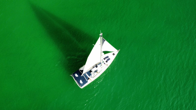 Aerial drone view of a white sailboat with a raised mainsail and navy cockpit cover, casting a long dark triangular shadow on vivid green water.