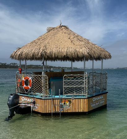 Floating tiki-hut bar with a thatched roof, rope-net railings, life preserver and outboard motor anchored in shallow clear turquoise coastal bay under a sunny sky