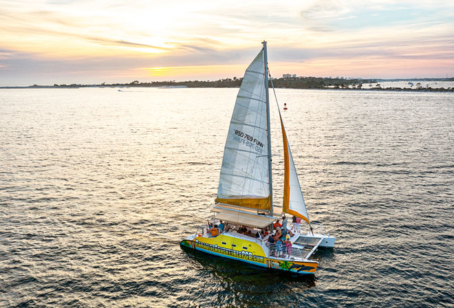 Vibrant yellow catamaran with tall white sails and passengers enjoying a sunset cruise across calm coastal waters near a low tree-lined shoreline.