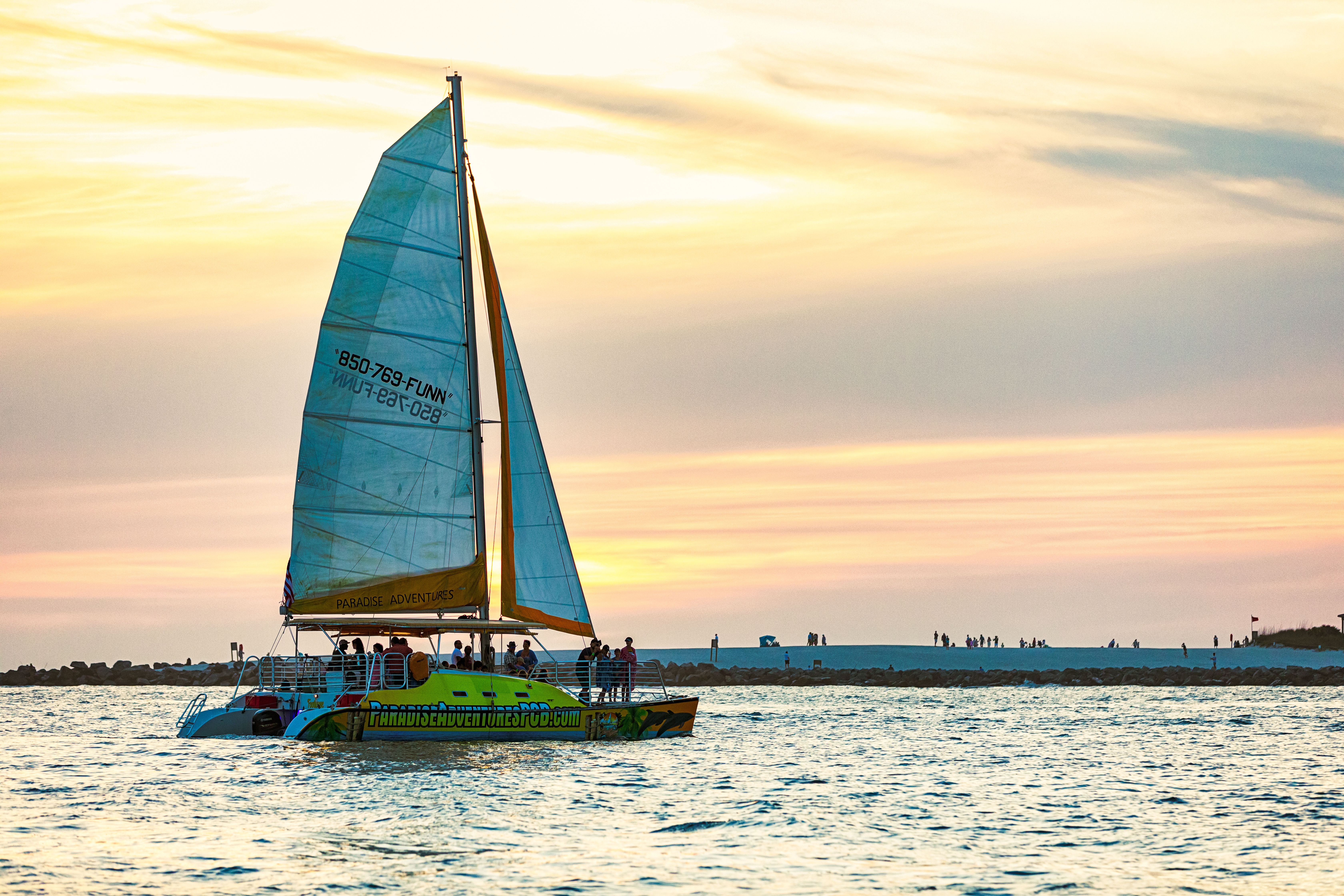 Colorful catamaran sailing at sunset near a sandy shoreline, passengers silhouetted on deck against a golden sky reflecting on calm ocean waters.