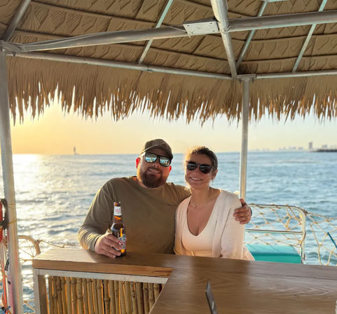 Smiling couple in sunglasses share a beer under a thatched tiki hut on a sunset ocean cruise, with a wooden bar and distant city skyline on the horizon.
