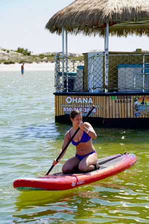 Woman in a blue bikini kneeling on a red paddleboard, paddling in shallow coastal waters near a thatched-roof floating tiki bar and sandy beach on a sunny summer day.