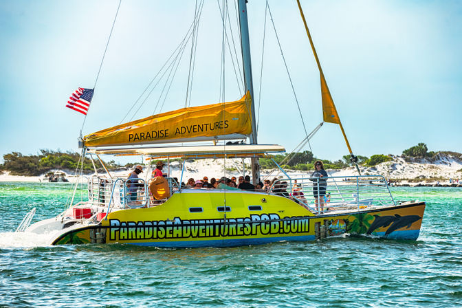 Bright yellow tour catamaran with passengers and an American flag cruising turquoise Gulf Coast waters past sandy dunes on a sunny day.
