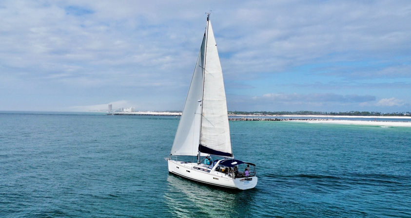 White sailboat with full mainsail gliding on turquoise coastal waters near a sandy shoreline under a partly cloudy blue sky