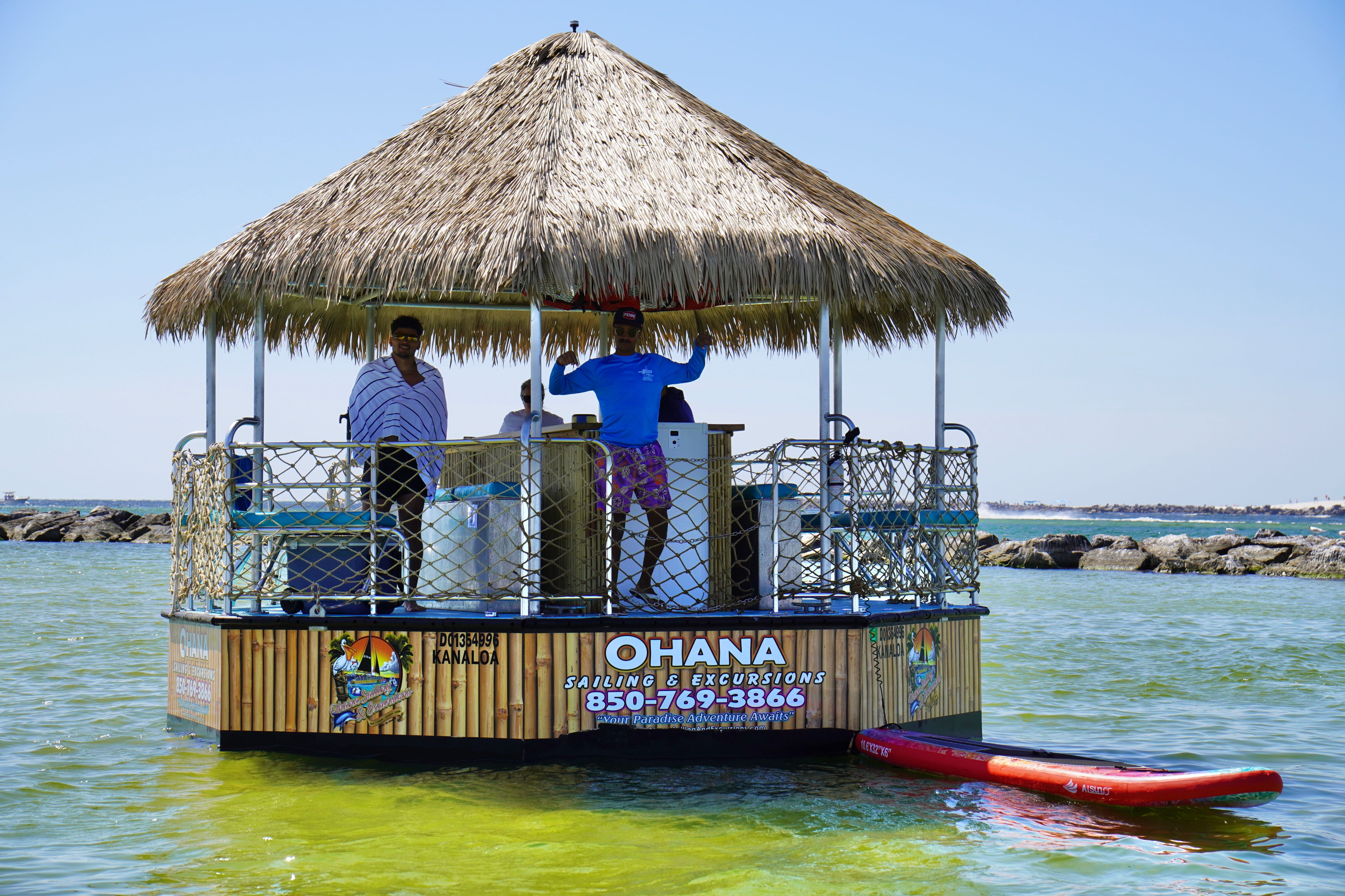 Thatched-roof floating tiki hut in clear shallow coastal waters, crew waving aboard a bay excursion vessel with a red paddleboard tied alongside and a rocky breakwater in the background.