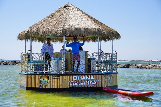 Thatched-roof floating tiki hut in clear shallow coastal waters, crew waving aboard a bay excursion vessel with a red paddleboard tied alongside and a rocky breakwater in the background.