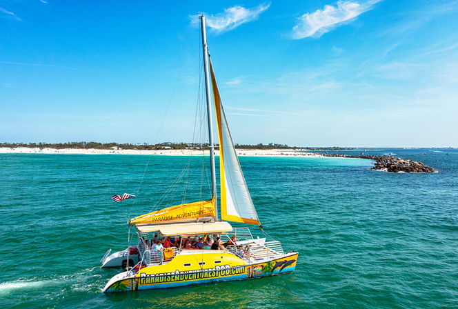 Bright yellow catamaran tour sailing on turquoise ocean past a white-sand beach and rocky jetty, passengers onboard under a sunny blue sky