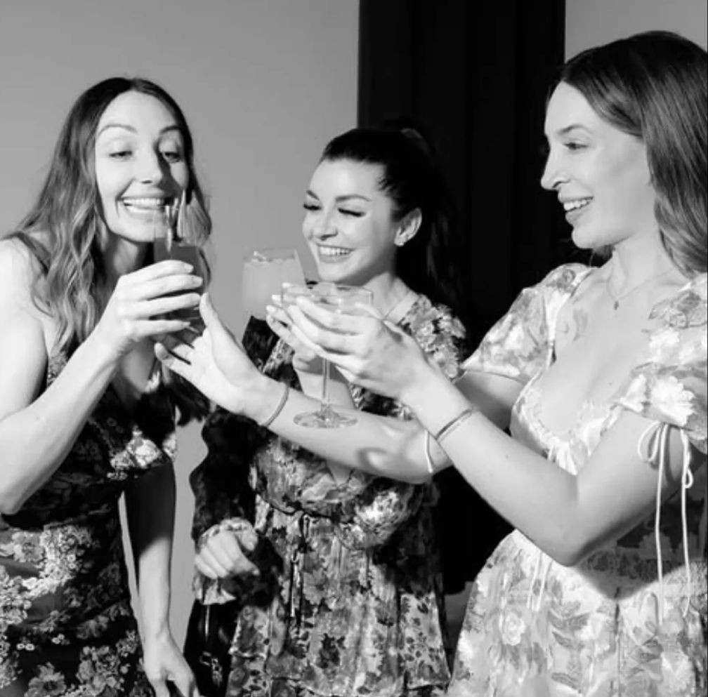 Three smiling women in floral dresses clinking cocktail glasses in a black-and-white indoor celebration photo.