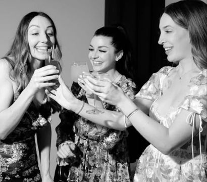 Three smiling women in floral dresses clinking cocktail glasses in a black-and-white indoor celebration photo.
