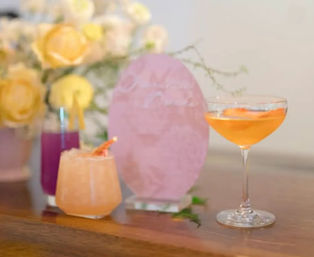 Three colorful cocktails—orange coupe, peach crushed-ice tumbler garnished with a peel, and a purple tall drink—arranged on a wooden bar with yellow roses and a pink decorative sign blurred in the background.