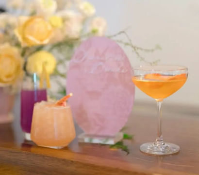 Three colorful cocktails—orange coupe, peach crushed-ice tumbler garnished with a peel, and a purple tall drink—arranged on a wooden bar with yellow roses and a pink decorative sign blurred in the background.