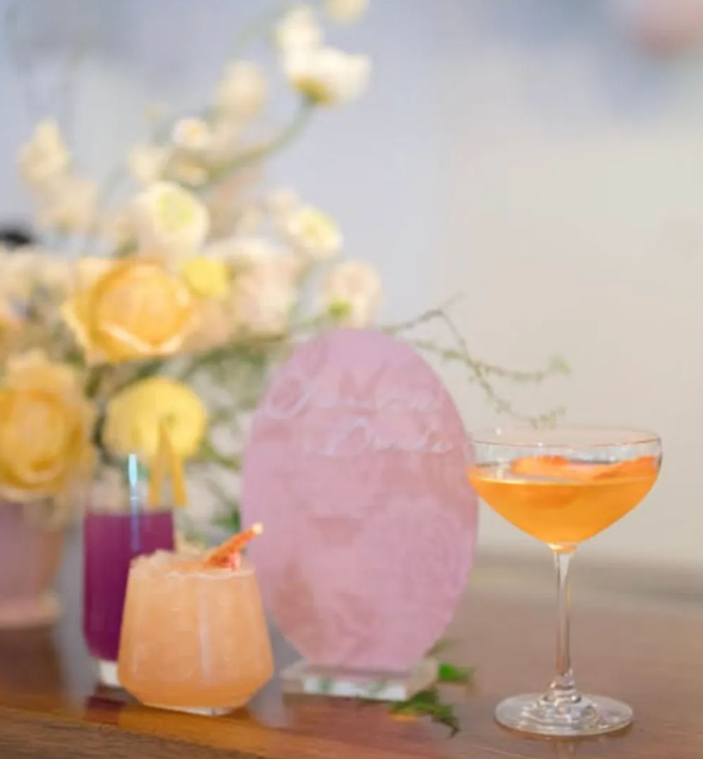 Three colorful cocktails on a wooden table — a crushed-ice peach lowball, a purple highball with straws and an orange coupe — arranged with a pink textured centerpiece and a soft-focus yellow rose bouquet in a pastel brunch setting.