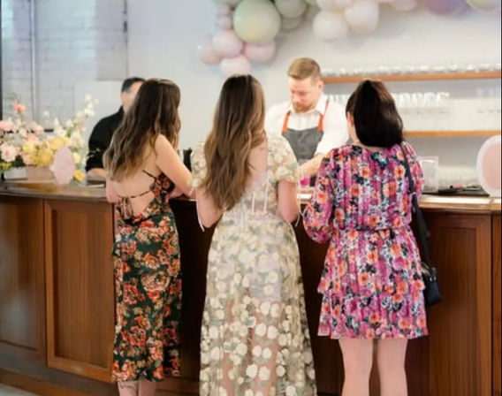 Three women in floral dresses chat with a bartender at a wooden bar inside a pastel-decorated event venue with balloons and flower arrangements.