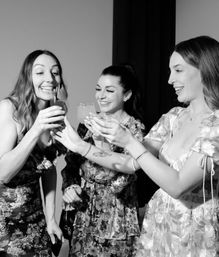 Three smiling friends in floral dresses clinking cocktail glasses at an indoor celebration — black-and-white, studio-style party photo.