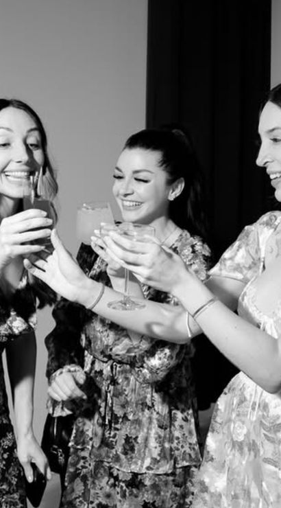 Three smiling friends in floral dresses clinking cocktail glasses at an indoor celebration — black-and-white, studio-style party photo.