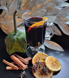 Warm mulled wine in a glass mug with an orange peel, surrounded by cinnamon sticks, star anise and dried orange slices beside a decorative green gourd and glittery silver leaves — festive autumn spiced drink.
