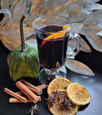 Warm mulled wine in a glass mug with an orange peel, surrounded by cinnamon sticks, star anise and dried orange slices beside a decorative green gourd and glittery silver leaves — festive autumn spiced drink.