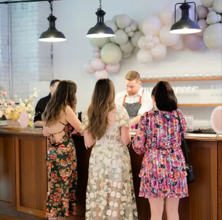 Three women in floral dresses at a wooden bar being served by a bartender under industrial pendant lights and a pastel balloon garland.